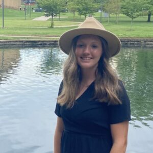 girl standing in front of water posing for an portrait wearing a hat and black dress