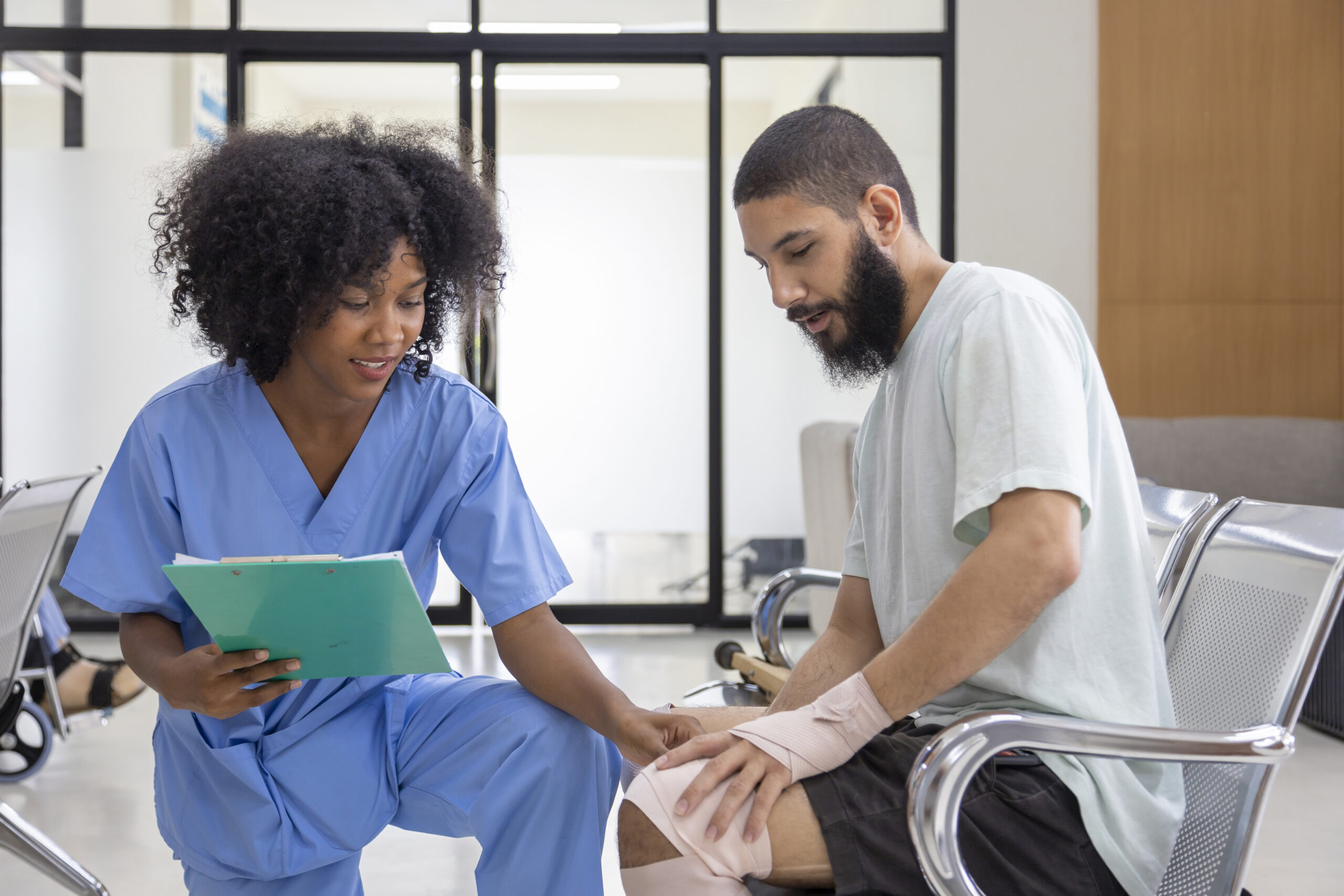 a black, female healthcare worker is kneeling in front of a young injured man with a beard in a hospital or medical setting.