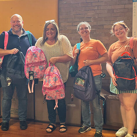 The team at Onward Injury Law stands with the school faculty inside the gymnasium holding up the backpacks they just donated to the school in Bloomington, IL.