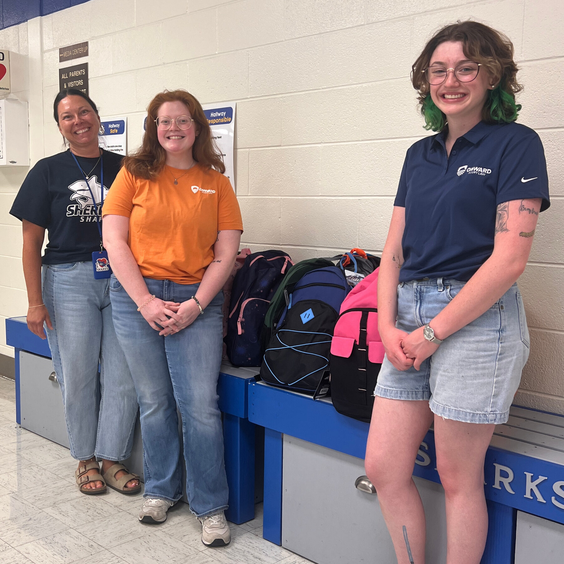two team members of Onward Injury Law standing with a school staff member in front of a group of backpacks they have donated to local students