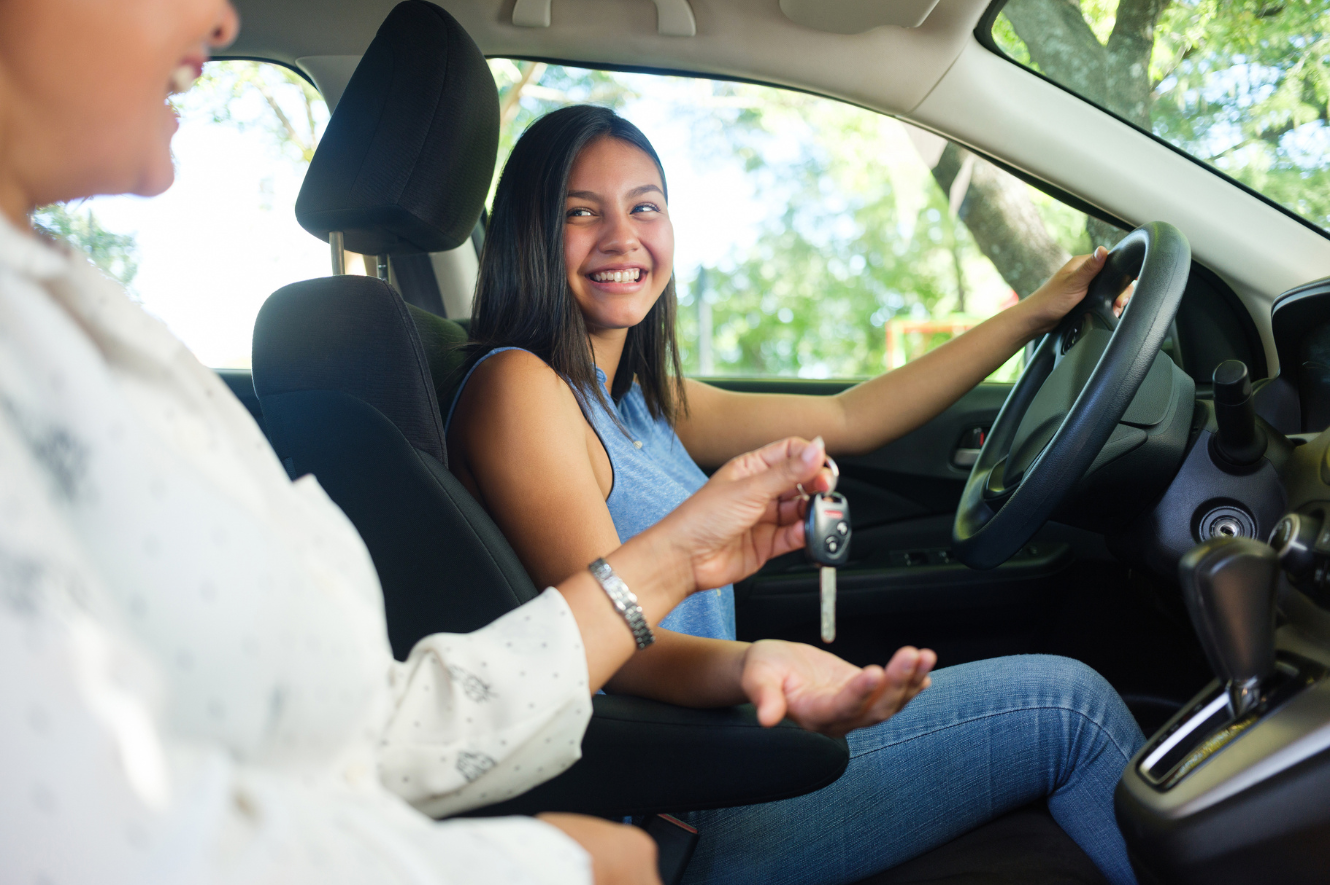 A side view photo of a teenage girl in a car as her mother sits next to her and hands her the keys to drive.